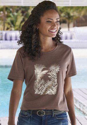 Helles, kurzes braunes T-Shirt mit einem weißen Palmenblattmuster, kombiniert mit blauen Jeans und einem geflochtenen schwarzen Gürtel mit einer runden Schnalle.