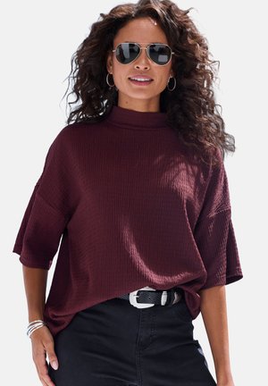 Frau mit lockigem Haar, die Pilotenbrille, Creolen, ein lockeres bordeauxrotes Oberteil, schwarze Hosen und silberne Armbänder trägt und selbstbewusst lächelt.