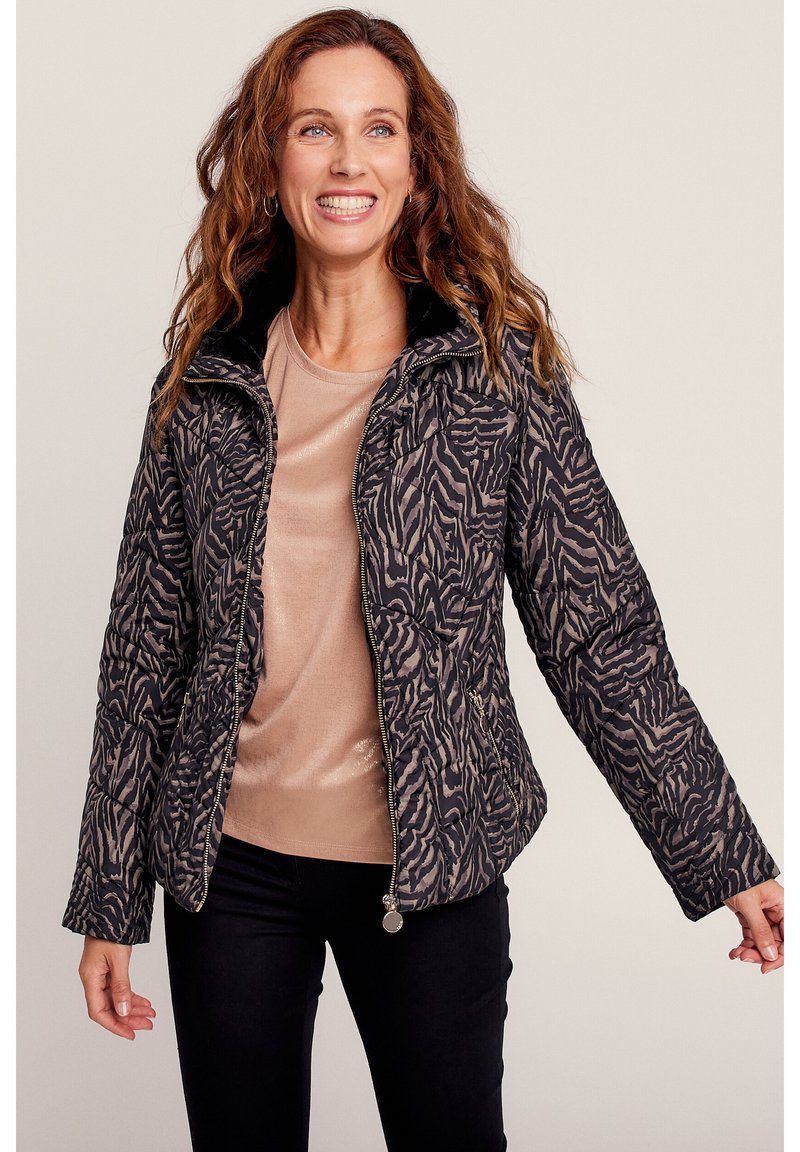 Ge quiltete Jacke mit einem schwarzen und beigen Zebra-Muster, hohem Kragen, Reißverschluss und Taschen an der Seite. Tragen über einem hellbeigen Oberteil.