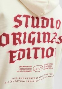 Béžová mikina s velkým červeným gotickým textem "Studio Originals Edition" a menším textem propagujícím městskou estetiku a každodenní oblečení.