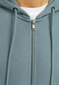Zip-hoodie u mekom tirkiznom materijalu, s srebrnim patentnim zatvaračem, kapuljačom s rastezljivom kosom i vidljivim šavovima. Glatka tekstura, ležeran dizajn.