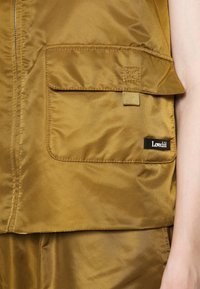 Satin vest in mustard yellow with a chest pocket, featuring a brand tag. Smooth texture with a zip closure and visible stitching details.