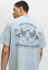 Lyseblå bomuld t-shirt med grafisk tryk af to vingede heste, krone og tekstelementer i marineblå. Korte ærmer og afslappet pasform.