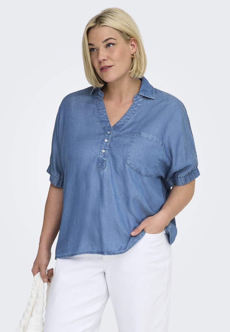 Femme blonde portant une chemise en jean ample et un pantalon blanc, tenant un sac blanc texturé, une main dans la poche, debout sur un fond uni.