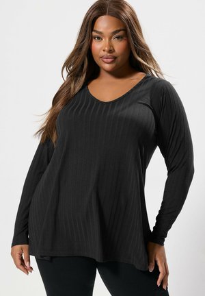 Frau mit langen braunen Haaren, die ein schwarzes, langärmliges, geripptes Oberteil und schwarze Hosen trägt, posiert vor einem schlichten hellen Hintergrund.