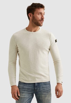 Man met kort bruin haar en baard, gekleed in een lichtbeige shirt met lange mouwen en blauwe spijkerbroek, kijkt naar zijn rechterkant.