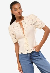 Pletený kardigán na zapínanie na gombíky v krémovej farbe s vlnitým vzorom, krátkymi nadýchanými rukávmi a čipkovými detailmi; v kombinácii s modrými džínsami s vysokým pásom.