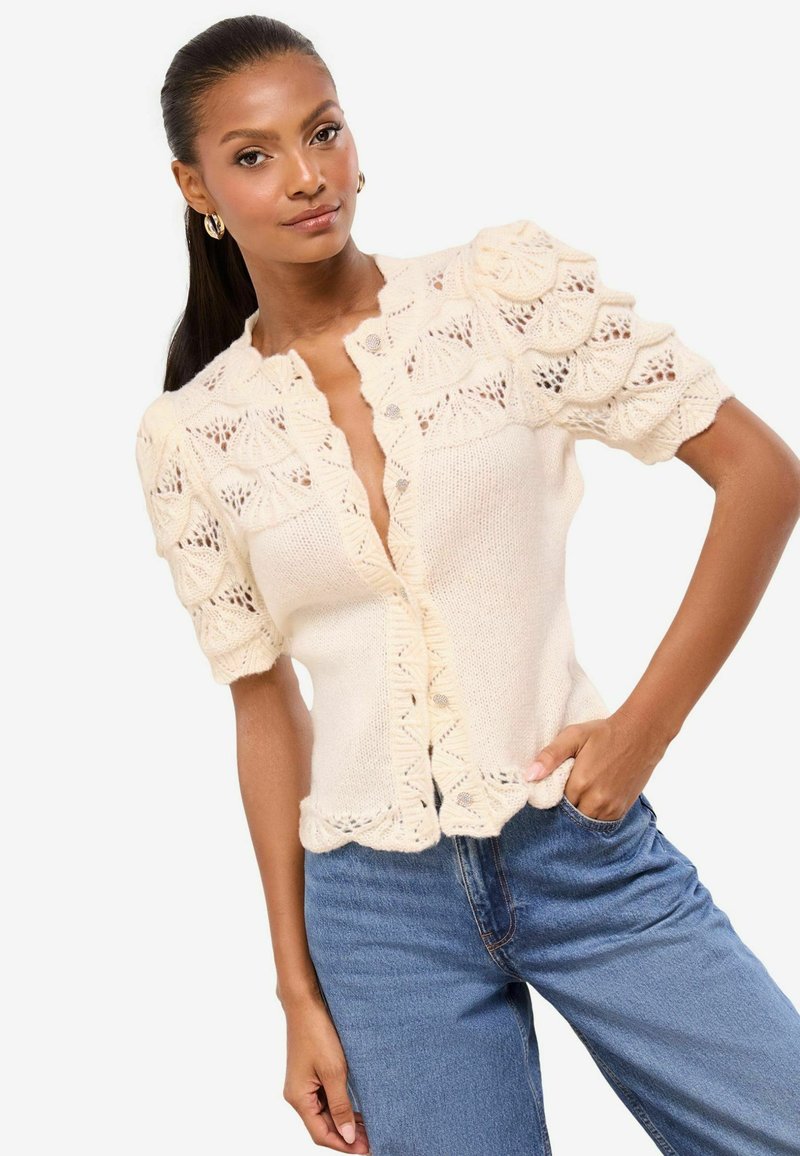 Pletený kardigán na zapínanie na gombíky v krémovej farbe s vlnitým vzorom, krátkymi nadýchanými rukávmi a čipkovými detailmi; v kombinácii s modrými džínsami s vysokým pásom.
