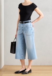 Žena v černém přiléhavém topu, světle modrých širokých zkrácených džínách s černým opaskem, černých mokasínech, držící černou kabelku, stojící na dřevěné podlaze.