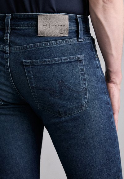 Temno modri denim kavbojki z gladko teksturo, ki imajo usnjen oznaki na pasu in šivane zadnje žepke. Opazni so subtilni oblikovni poudarki.