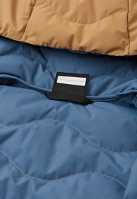 Tessuto blu trapuntato con uno strato superiore beige, dotato di un anello nero per etichette. Texture liscia e motivi di cucitura ondulati a contrasto visibili.