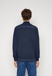 Man med lockigt brunt hår iförd en marinblå tröja och blå jeans, stående vänd mot en enkel ljus bakgrund.