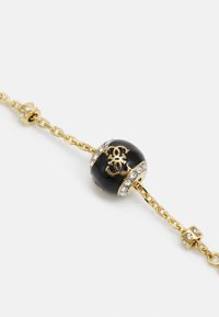 Bracelet en or avec une perle noire ronde portant un logo en or, accentué par de petits cristaux clair le long de la chaîne et des bords de la perle.