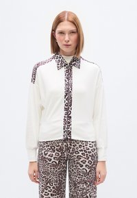 Λευκό πουλόβερ με λεοπάρ λεπτομέρειες στο γιακά και στο μπροστινό μέρος, με μακριά μανίκι α και άνετη γραμμή. Συνδυάζεται με παντελόνι σε λεοπάρ σχέδιο.