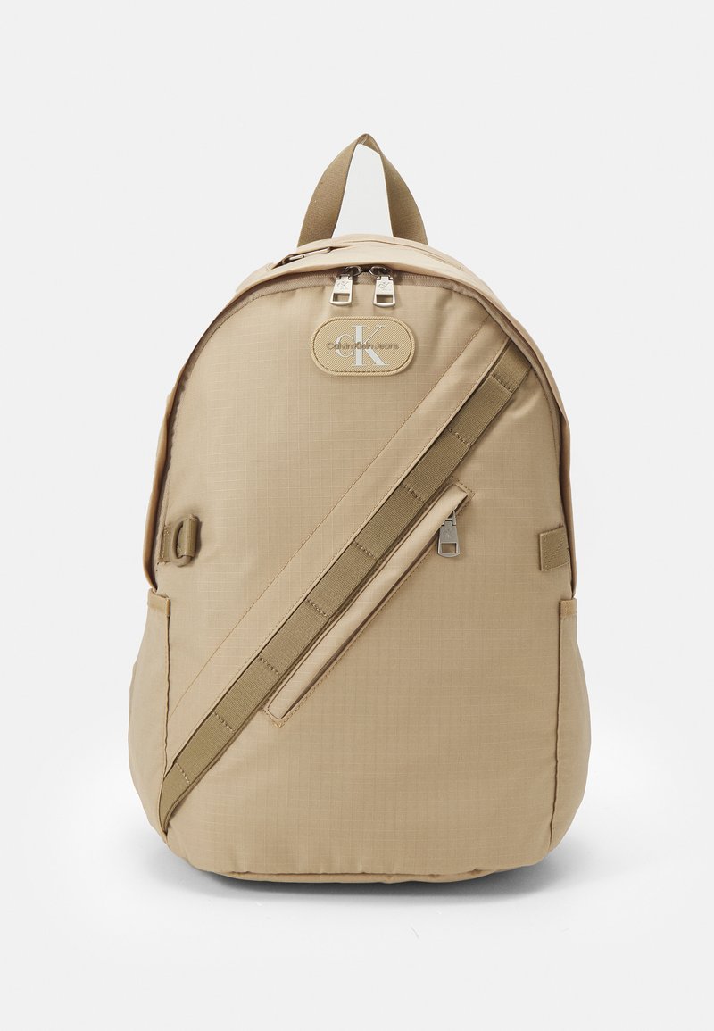 Zaino beige, realizzato in tessuto resistente, presenta una parte superiore arrotondata, una tasca frontale con zip, una patch con il logo e spallacci regolabili per il massimo comfort.