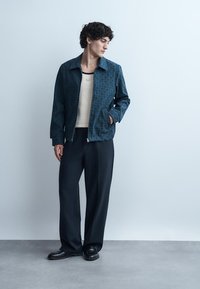 Moški s kodrastimi lasmi stoji obrnjeno desno, oblečen v temno vzorčasto jakno, bež srajco, ohlapne črne hlače in črne čevlje, obdan s svetlim zidom.