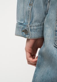 Χέρι χαλαρό στο πλάι με ελαφρύ μπλε τζιν μπουφάν, με ορατό κουμπί στο μανίκι και λεπτομέρειες ραφής.