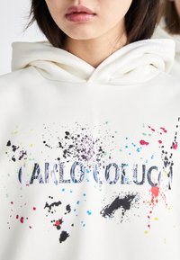 Hoodie blanc avec un graphique sur le devant comportant le texte "CARLO COLUCCI" et des motifs de taches colorées en noir, bleu, vert et rouge.