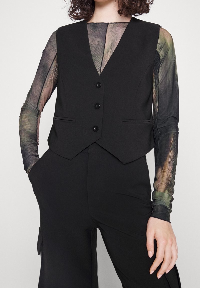 Femme portant un gilet noir boutonné sur un top à manches longues transparent à motifs avec un pantalon noir, mains dans les poches sur un fond uni.