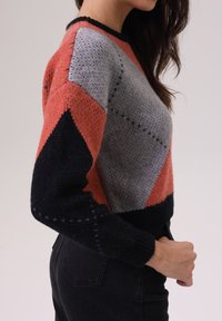 Pull en tricot avec un motif géométrique dans des tons d'orange, de gris et de noir. Design cropped avec des poignets et un ourlet côtelés.