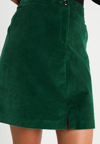 Gonna in velluto a coste verde con righe verticali, spacco frontale e vita alta con dettaglio a bottone. Texture morbida e vestibilità strutturata.