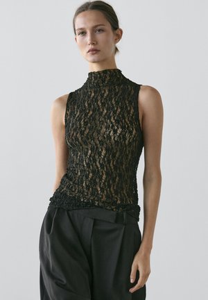 Femme portant un haut sans manches en dentelle noire à col haut et un pantalon noir ample, debout devant un fond clair uni.