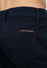 Pantaloni blu navy con una texture liscia, caratterizzati da dettagli di cucitura rossa e un'etichetta rettangolare con la scritta "CIPO & BAXX" sulla tasca posteriore.