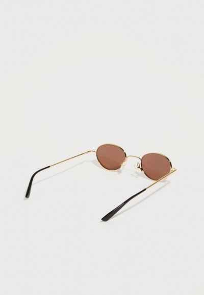 Στρογγυλά καφέ φακοί γυαλιών ηλίου με λεπτό χρυσό μεταλλικό πλαίσιο και μαύρες άκρες στα βραχίονες, σε λευκό φόντο.