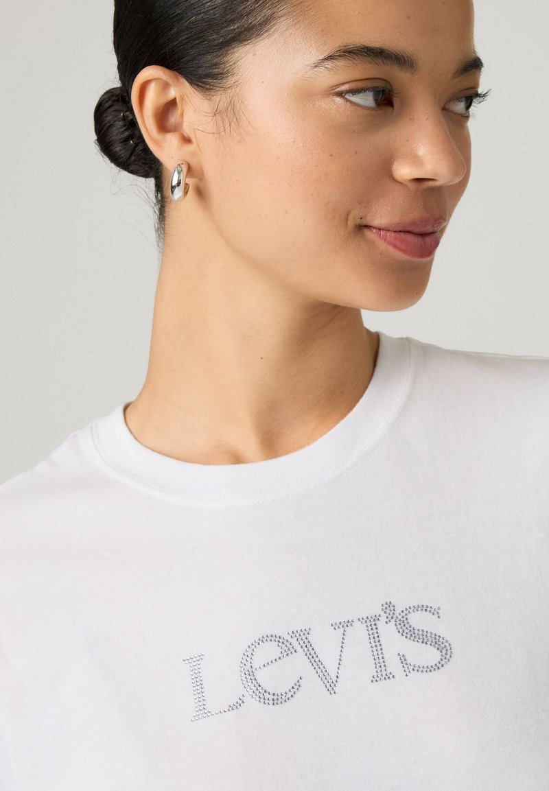 Femme aux cheveux foncés attachés en chignon, portant des boucles d'oreilles créoles argentées et un t-shirt blanc Levi's avec un logo en strass, regardant sur le côté et souriant légèrement.