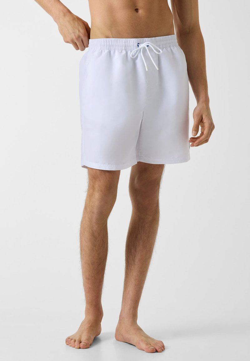 Personne pieds nus portant un short blanc à taille élastique avec cordon, debout devant un fond clair uni, ajustant la taille d'une main.