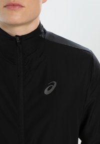 Schwarze Zip-Jacke mit hochgeschlossenem Kragen, aus leichtem Material gefertigt. Verfügt über ein reflektierendes Logo auf der Brust und eine glatte Textur.
