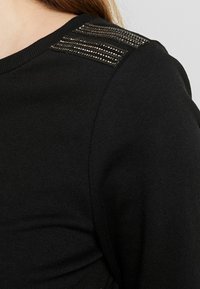 Pull noir en tissu texturé avec des rayures décoratives en sequins sur l'épaule. Vue en gros plan mettant en valeur le détail de l'épaule.