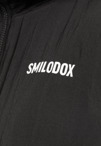 Czarna kurtka z teksturowanym wykończeniem, z logo "SMILODOX" wytłoczonym białymi literami po lewej stronie. Zapięcie na zamek.