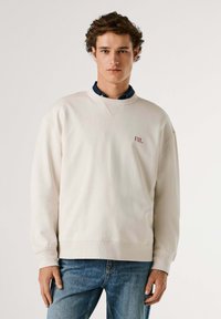 Felpa color crema con scollatura rotonda, polsini e orlo a costine. Presenta un piccolo logo rosso "PJL" sul petto. Indossata sopra una camicia blu con colletto.