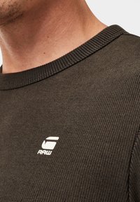 Donkergroene geribbelde sweatshirt met een ronde hals en een klein wit logo op de borst. De stof heeft een gestructureerde uitstraling.