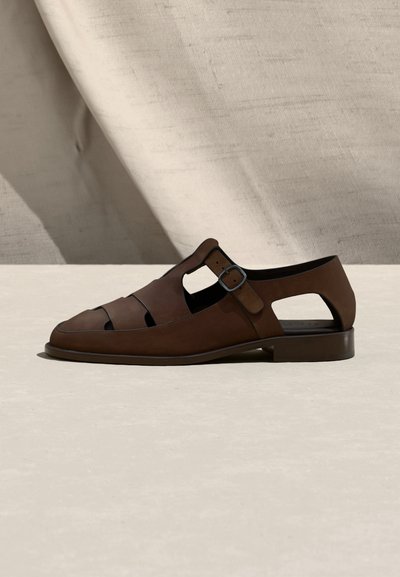 Καφές δερμάτινος ανδρικός σανδάλι με ανοίγματα και αγκράφα στο πλάι, τοποθετημένο σε ουδέτερο ύφασμα φόντο και ανοιχτόχρωμη επιφάνεια.