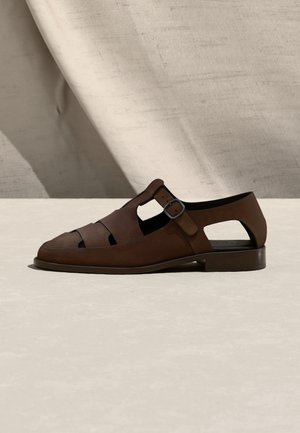 Muške smeđe kožne sandale s izrezima i bočnom kopčom, postavljene na neutralnu tkaninu i svijetlu površinu.