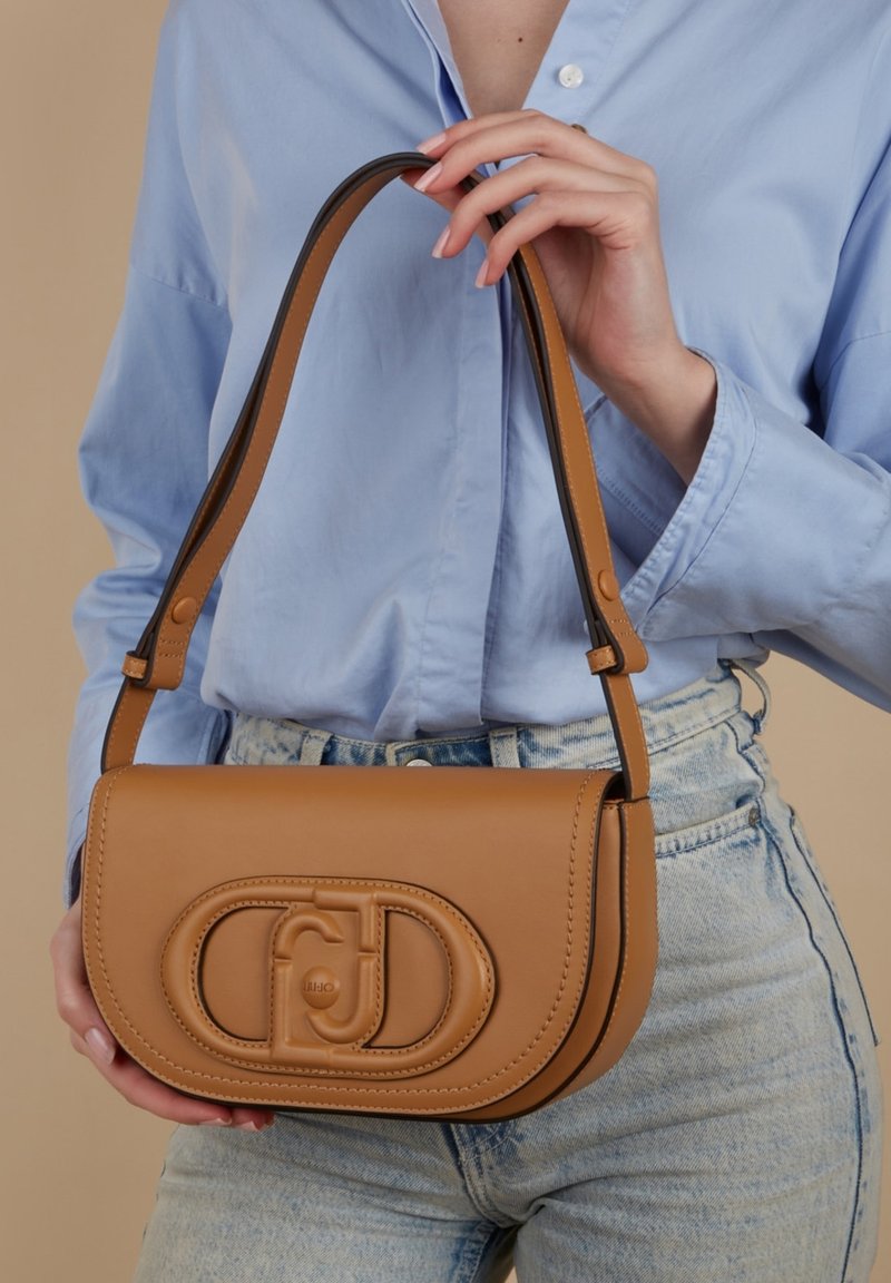 Bolso de hombro de cuero en tono tostado con forma curvada, que presenta un destacado detalle del logotipo y una textura suave. La correa tiene acentos en negro contrastantes.