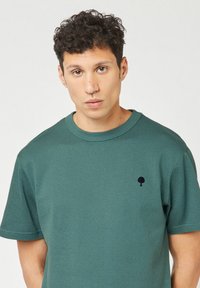 Man met kort krullend haar die een groen textuur korthangend shirt draagt met een klein zwart boomlogo op de borst, met het gezicht naar voren.