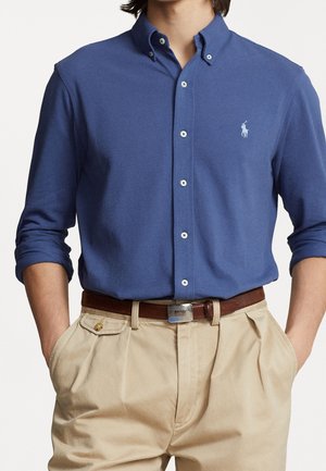 Mann trägt ein blaues Hemd mit Knöpfen und Polo-Logo, beige plissierte Hose, brauner Gürtel und Hände in den Taschen.