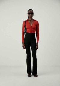 Vrouw in zwarte zonnebril, rood strakke blouse met knopen, hoge zwarte broek, zwarte handtas en zwarte sandalen die tegen een effen achtergrond staat.