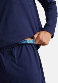 Top de manga larga azul marino con un bolsillo, combinado con pantalones a juego que tienen un forro interior a cuadros en azul y verde visible en la cintura.