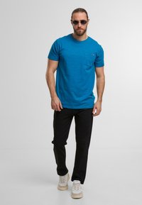 Blaues Kurzarm-T-Shirt mit Brusttasche und strukturiertem Stoff, kombiniert mit schwarzen Hosen und hellen Sneakers.