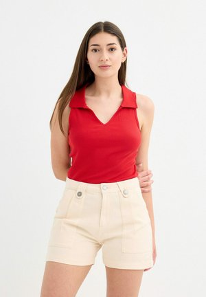 Top sin mangas de color rojo con canalé y cuello, combinado con pantalones cortos de talle alto en color beige que cuentan con dos bolsillos frontales y botones decorativos.