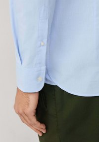 Camisa de botones a cuadros en azul claro con un puño doblado, que presenta un dobladillo redondeado y un pantalón de color verde oscuro en contraste. Los botones son perlados.