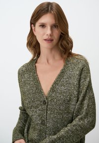 Groene gebreide cardigan met een V-hals, twee bruine knopen en een gestructureerd patroon. Zacht, relaxte pasvorm met iets taps toelopende mouwen.