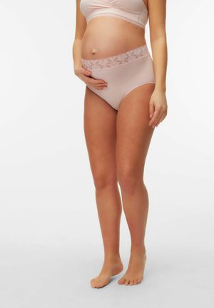 Femme enceinte en sous-vêtements de maternité rose clair ornés de dentelle, tenant doucement son ventre, debout pieds nus sur un fond blanc.