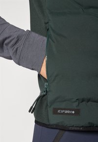 Giacca a vento verde con tasca con zip e dettagli con lacci a toggle. Sotto presenta una maglietta grigia a maniche lunghe. Branding visibile sull'orlo.