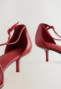 Zapatos de tacón alto de cuero rojo con un tacón fino y correa al tobillo. Textura suave, parte trasera abierta y una plantilla de marca visible.