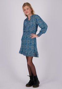 Junge Frau mit blonden Haaren posiert in einem blauen gemusterten Kleid, schwarzen Netzstrumpfhosen und schwarzen Schnürstiefeln vor einem schlichten Hintergrund.